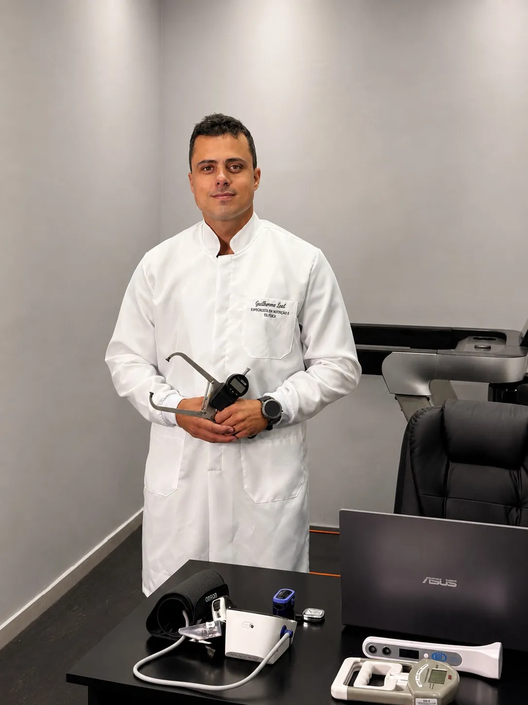 Guilherme Leal de jaleco na sala de avaliação, com o adipômetro na mão e os aparelhos da avaliação sobre a mesa